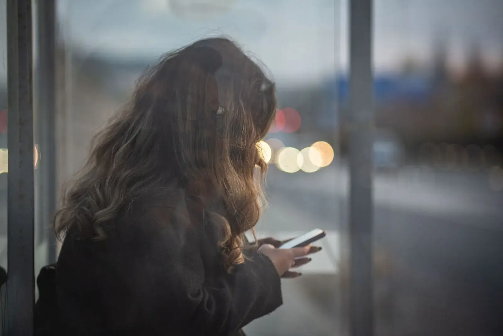 A woman checks her smartphone in a glass bus shelter at dusk, with blurred traffic lights behind her.