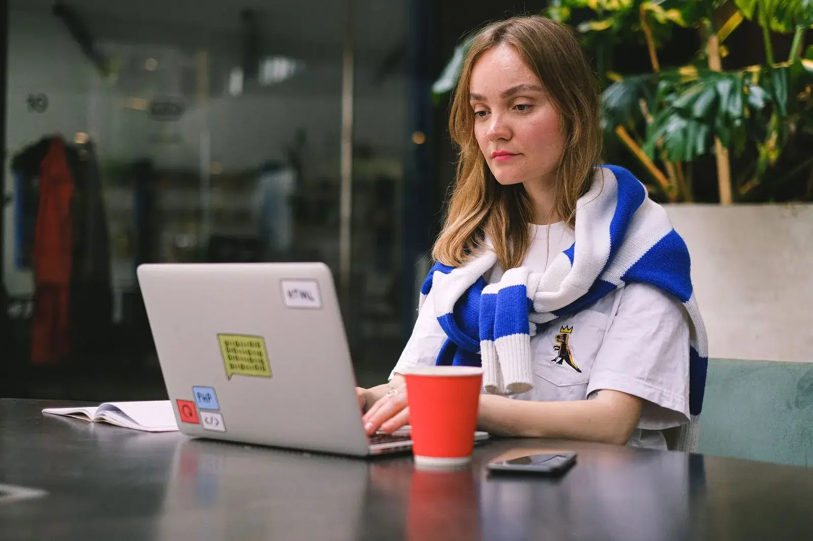 A web developer works on a laptop at a café table, with her phone on the table beside it.