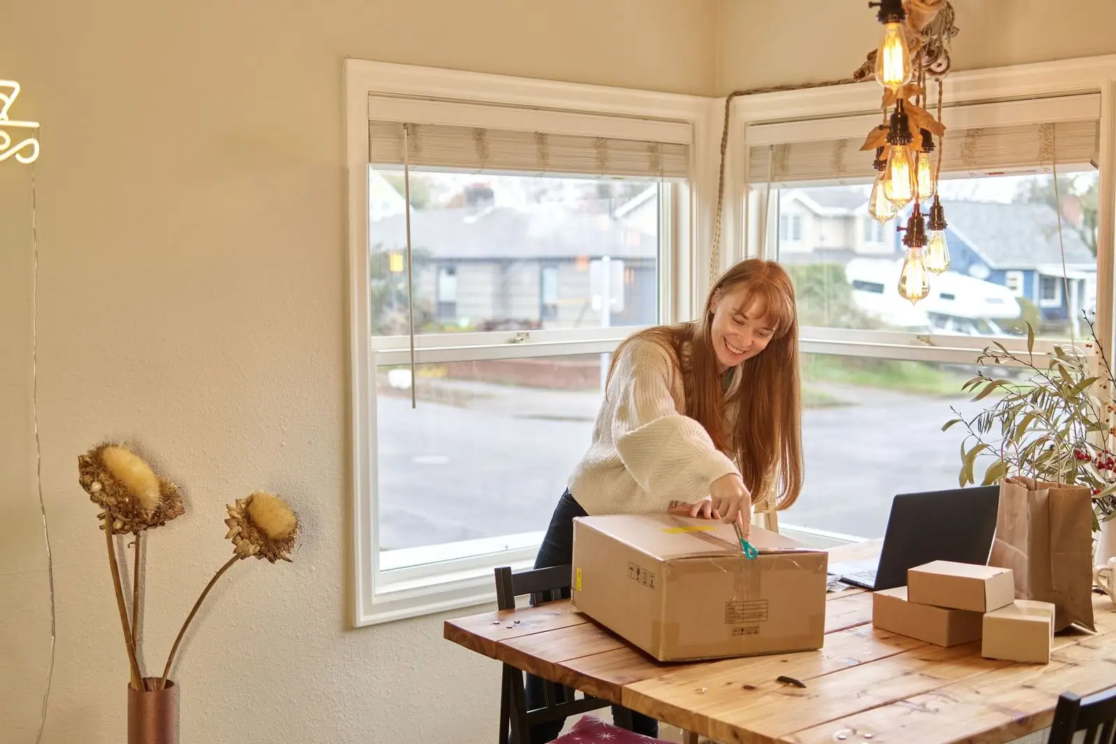 A woman at a wooden table opening a cardboard delivery box in a bright home.